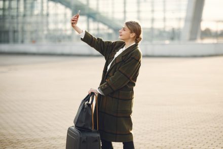 A woman in a plaid coat takes a selfie while holding a suitcase